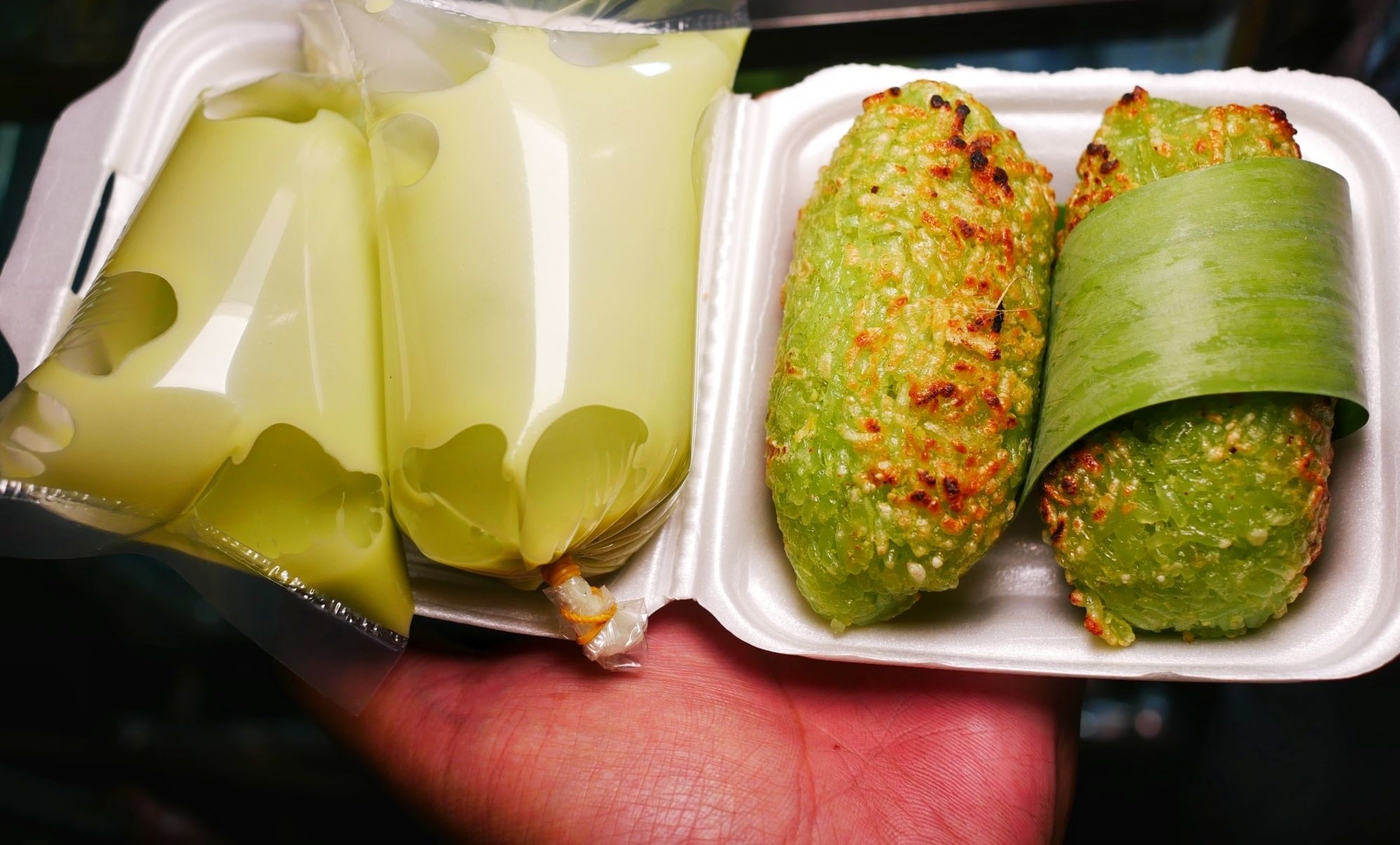 Grilled banana wrapped in white sticky rice, charred slightly from the grill, served with coconut milk sauce